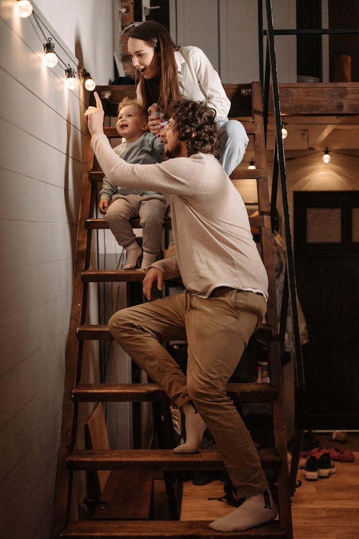A family enjoys quality time indoors, sitting on wooden stairs lit by string lights.